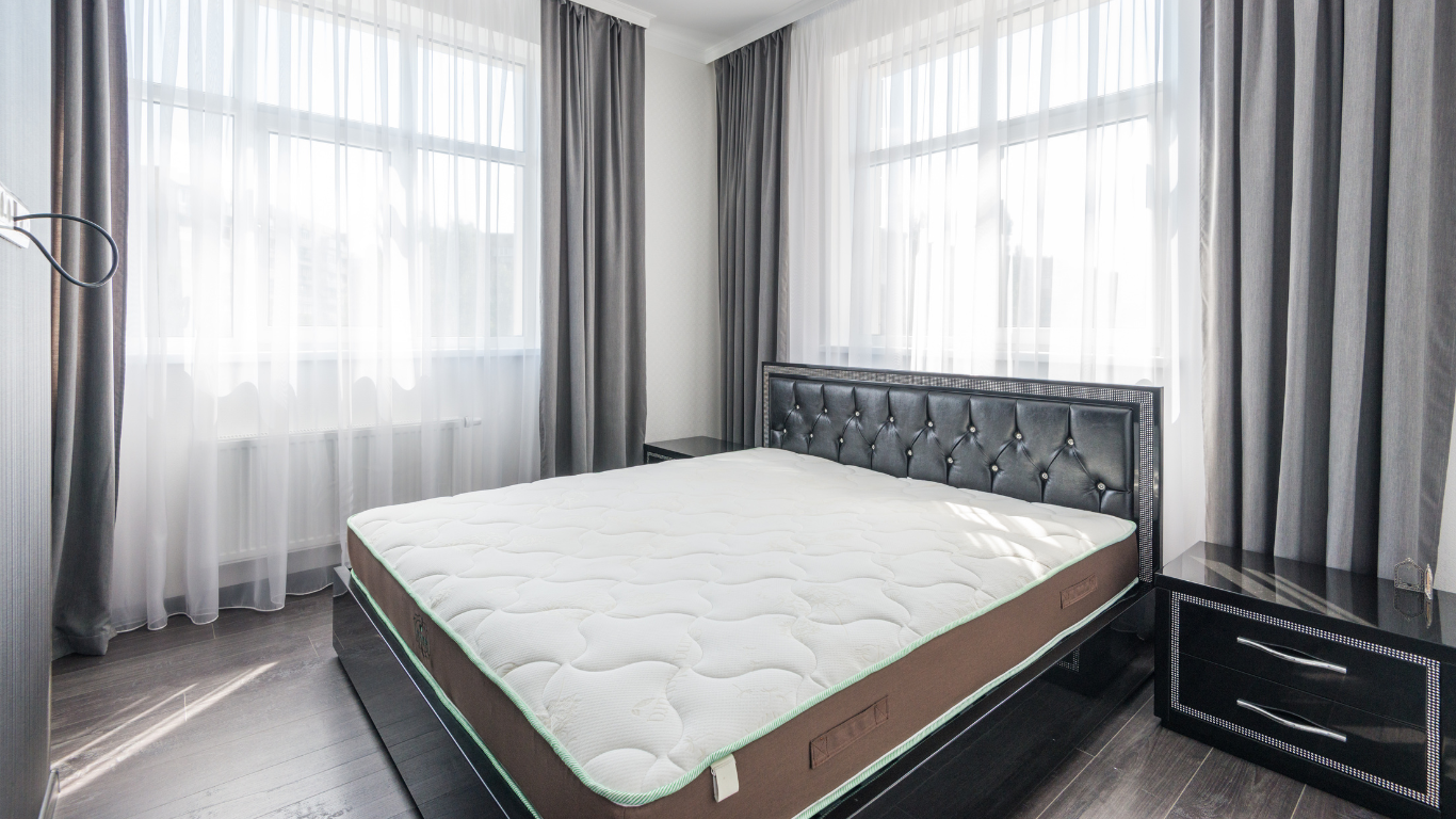 Room with natural light, large windows and a bed with an uncovered mattress.
