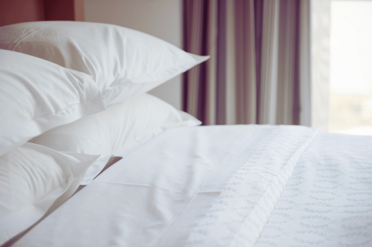 closeup of an all white linen bed with four pillows and curtains in the background