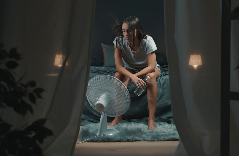 woman sitting up in bed sweating with a fan on her