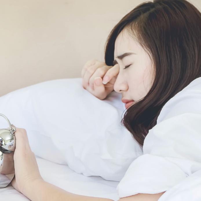 Young woman with one hand on alarm clock while rubbing her eye upon awakening