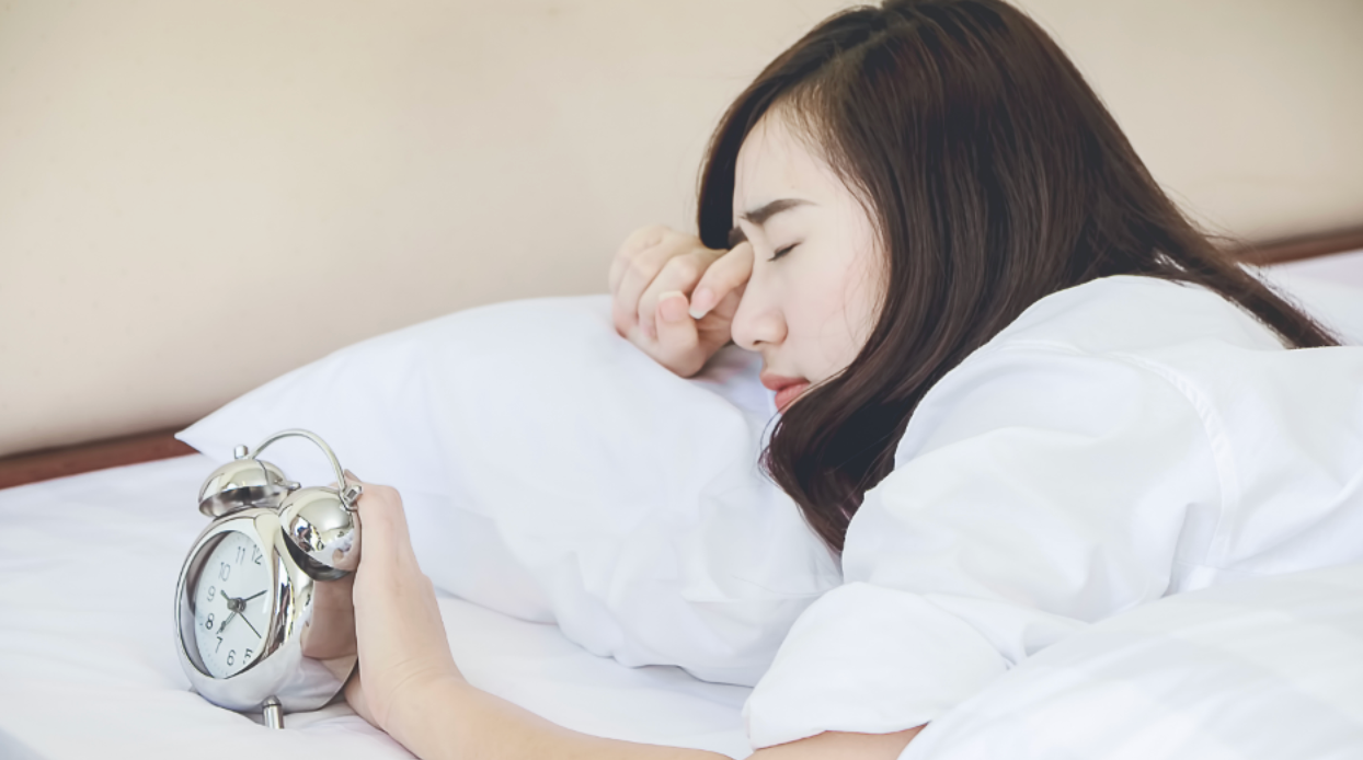 Young woman with one hand on alarm clock while rubbing her eye upon awakening
